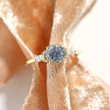 A stormy gray-blue sapphire engagement ring with 6 accent diamonds set in 14k yellow gold, shown on peach velvet fabric.